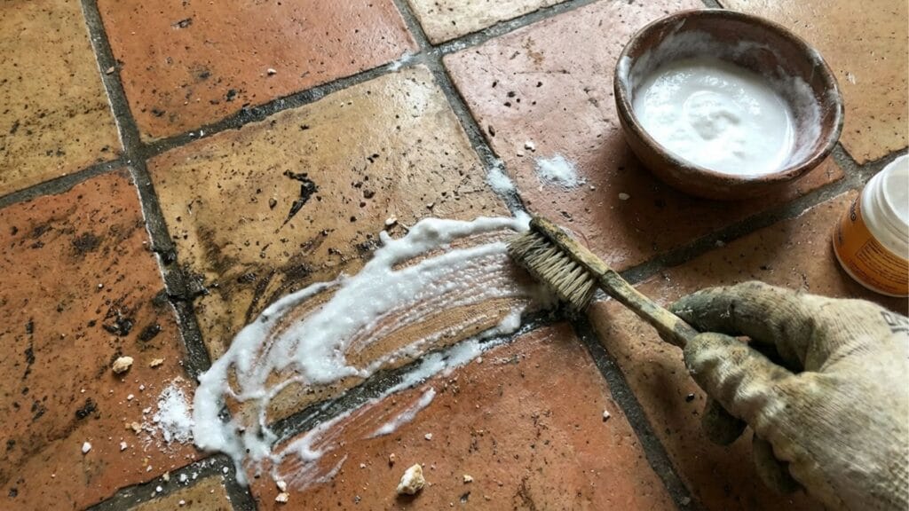 Reinigung von schmutzigen Fliesenfugen auf einem Terrakotta-Boden mit einer Zahnbürste und Natron-Paste.